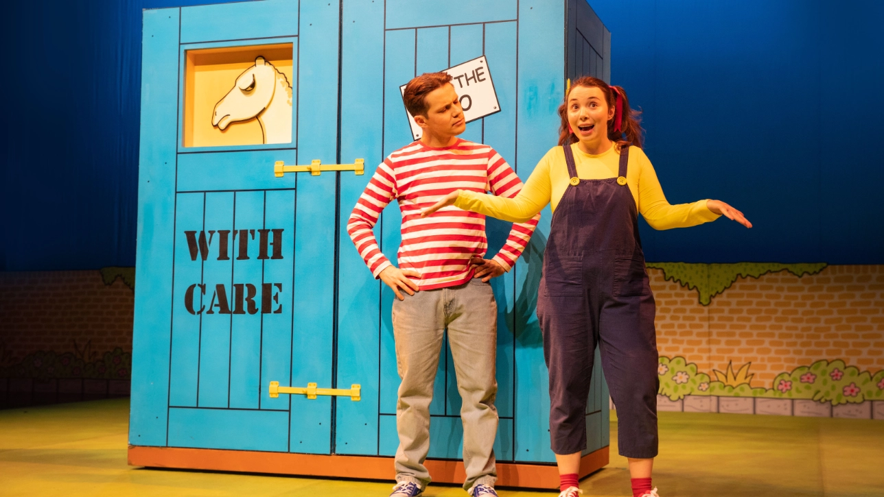 Two playful performers pose with a large green crate labeled “fragile” and “from the zoo,” from which a cartoon giraffe head emerges. The scene is colorful and whimsical.