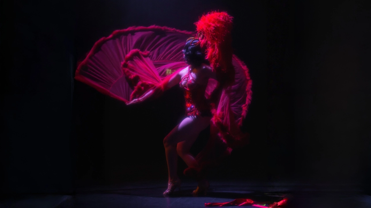 A performer in a dynamic pose is dressed in an elaborate red costume with large feathers, set against a dark background, conveying boldness and drama.