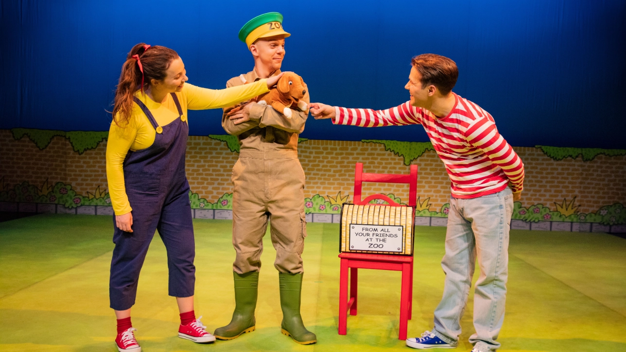 Three performers are onstage in a colorful setting. A person in a greenlds a  zoo uniform hoplush animal, as a woman and man touch it playfully. A red chair is nearby.
