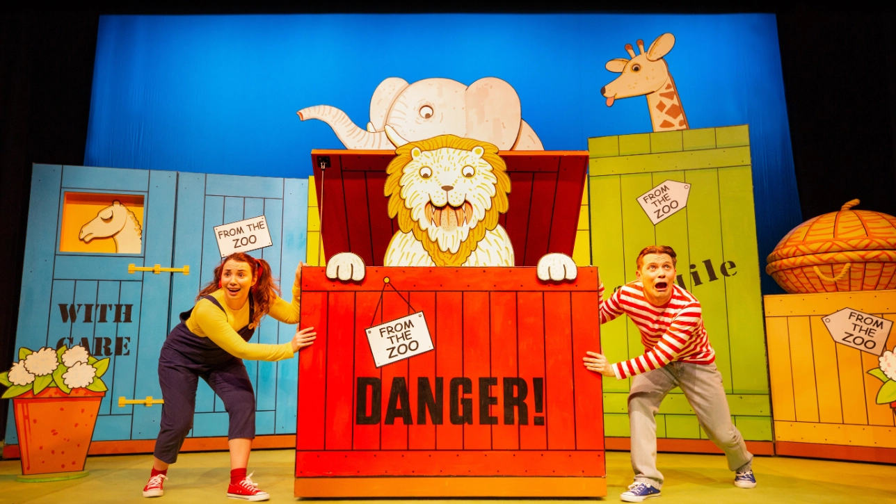 Two performers in colorful attire pose theatrically on stage with large, cartoon-style zoo crates. A lion, elephant, and giraffe are illustrated.
