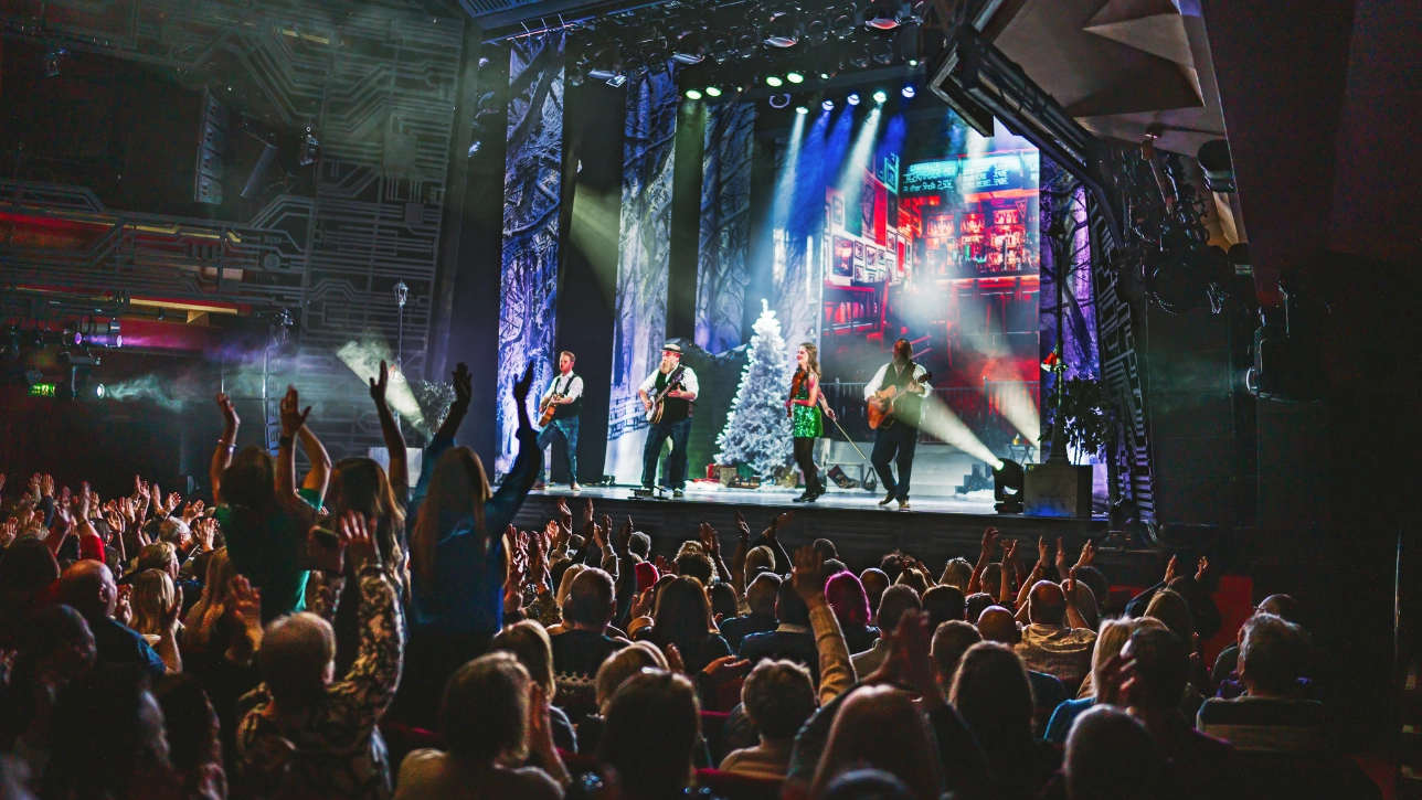  lively theatre audience cheers and claps as a band performs on stage. The musicians, dressed in festive attire, include a guitarist, a female singer in a green dress, and other instrumentalists. The stage design combines wintery elements, including snow-covered trees, with a backdrop of an Irish pub. Bright stage lighting illuminates the performers while silhouettes of enthusiastic audience members raise their hands in the foreground.