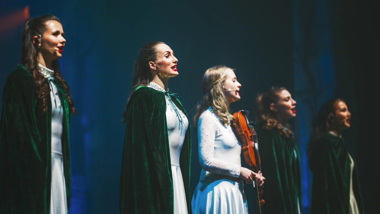 A group of female vocalists and musicians perform on stage, dressed in elegant white dresses with green velvet capes. One singer, standing at the front, sings passionately with red lips and expressive eyes, while another musician holds a violin. The stage lighting casts a soft glow on their faces, creating an ethereal and enchanting atmosphere.