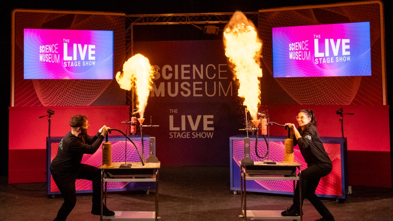 Two presenters on stage at the Science Museum's Live Stage Show demonstrate an explosive experiment, creating two tall flames shooting into the air. They are both wearing black uniforms and safety glasses, engaging the audience with an exciting fire demonstration. The background features large screens displaying the show's branding in pink and blue.