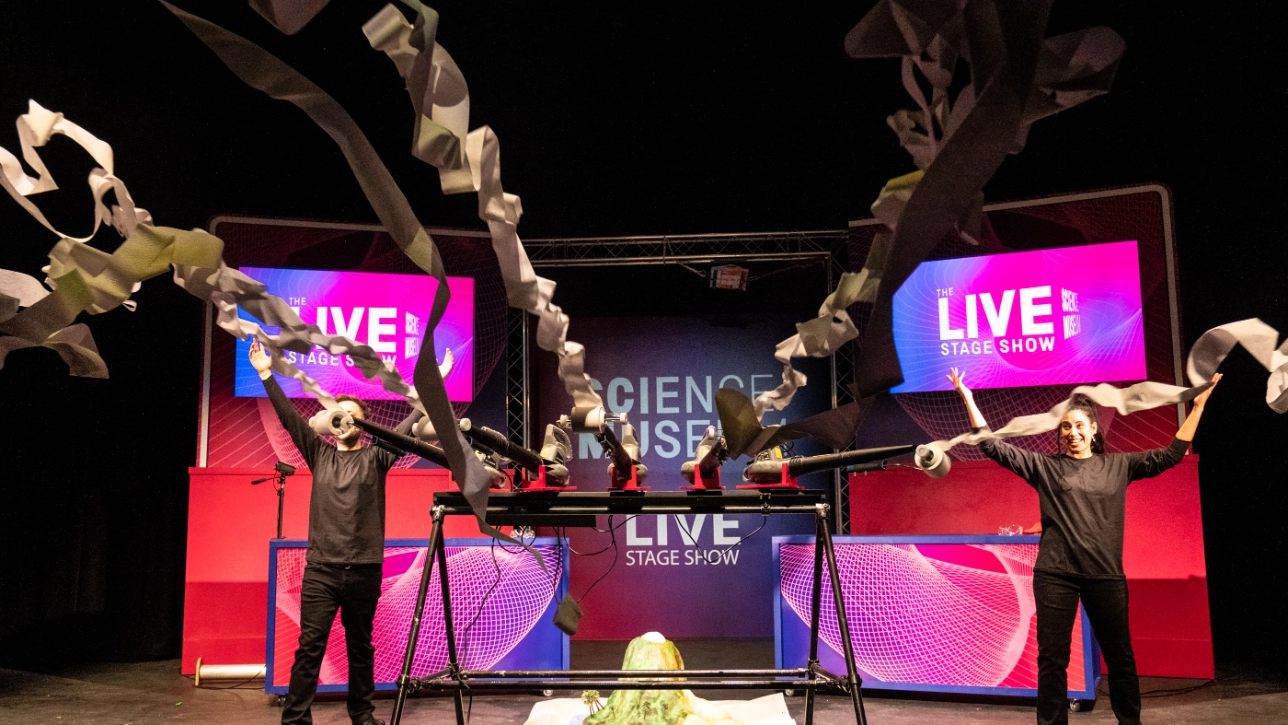Two presenters on stage at the Science Museum’s Live Stage Show perform an energetic experiment, launching long streams of toilet paper into the air using leaf blowers. A foamy volcano-like reaction is visible on the table, with the vibrant stage setup and branding displayed in the background.