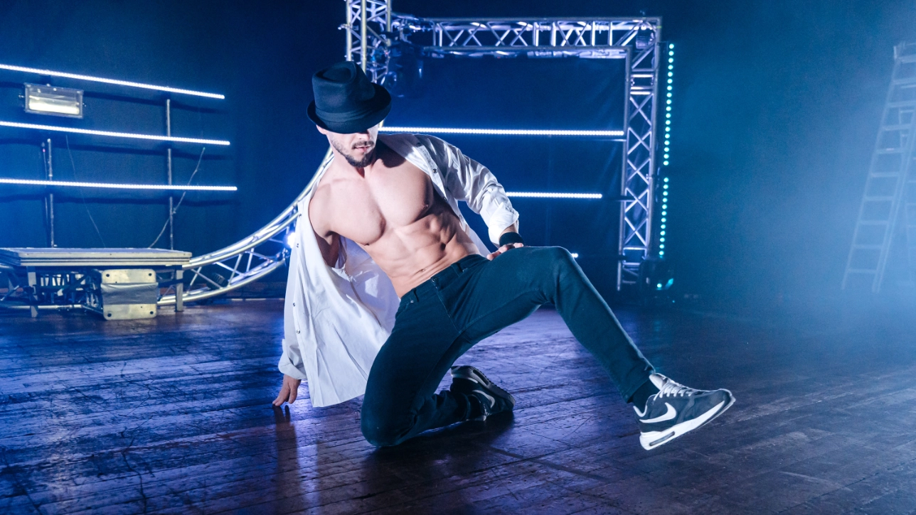 A shirtless male dancer wearing an open white shirt, black pants, sneakers, and a black fedora performs a floor move on a stage.
