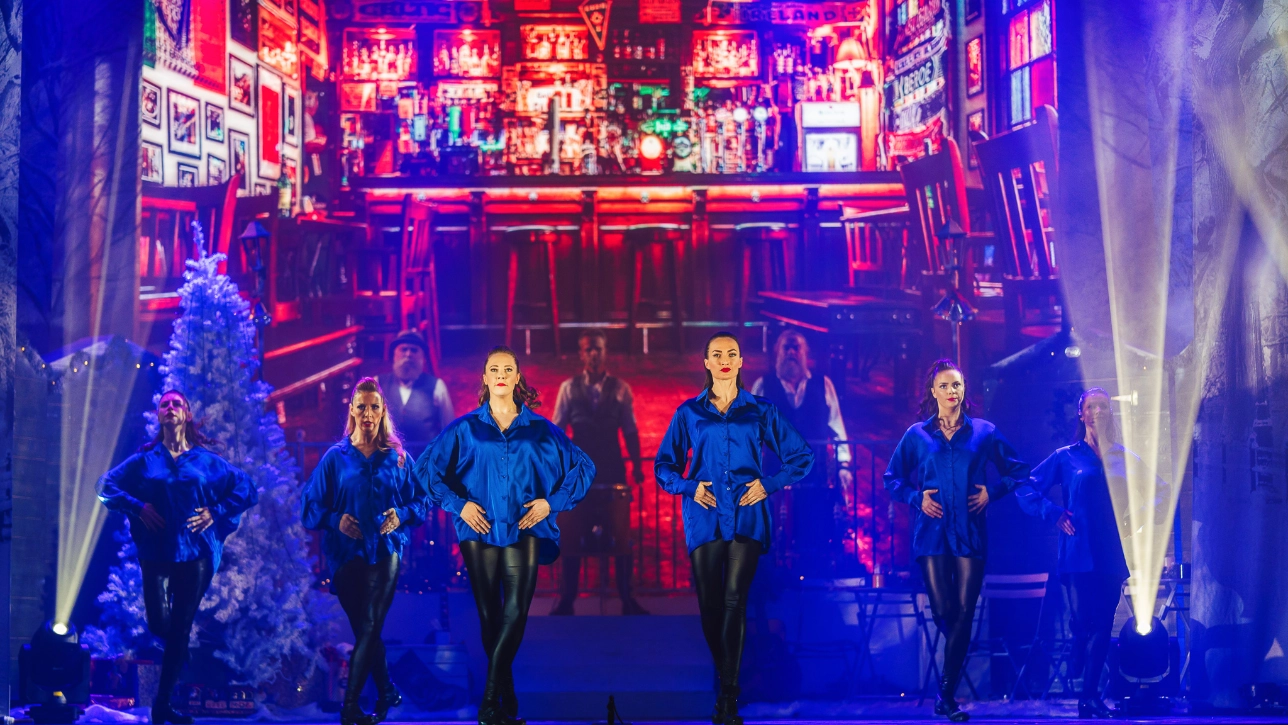 A group of Irish dancers performs on stage, wearing matching royal blue satin blouses and black leggings. They are mid-step, with hands on their waists and feet poised. The backdrop features a vibrant Irish pub scene with glowing red and orange lighting, creating a warm contrast to the cool-toned costumes. The stage is decorated with a snow-covered Christmas tree, and atmospheric lighting enhances the performance.