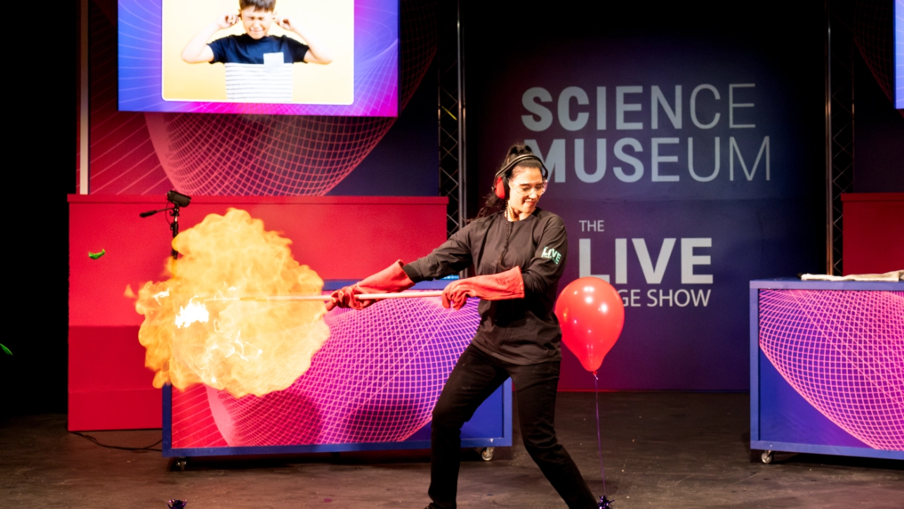 A presenter in a black uniform and safety gear, including ear protection and red gloves, holds a flaming torch at the Science Museum’s Live Stage Show. A red balloon is tied to their foot, and a screen in the background shows an image of a child covering their ears, hinting at a loud or explosive demonstration.