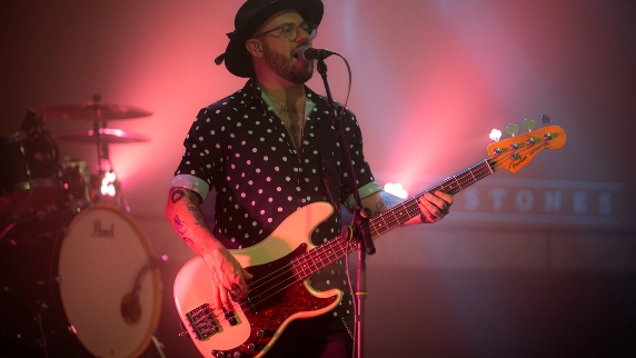 A musician wearing a hat and glasses plays an electric bass guitar and sings into a microphone on stage with pink and purple lighting.
