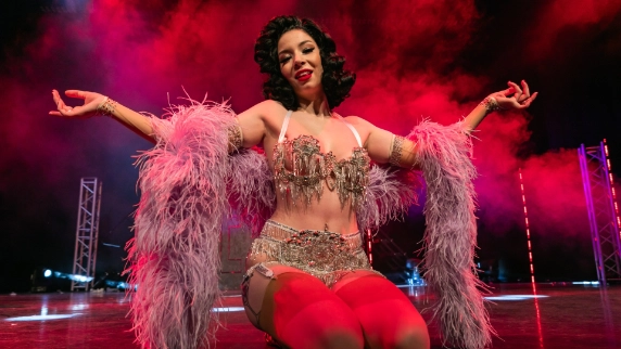 Performer in an embellished outfit and feather boa kneels on stage, enveloped in red smoke. The atmosphere is vibrant and theatrical.