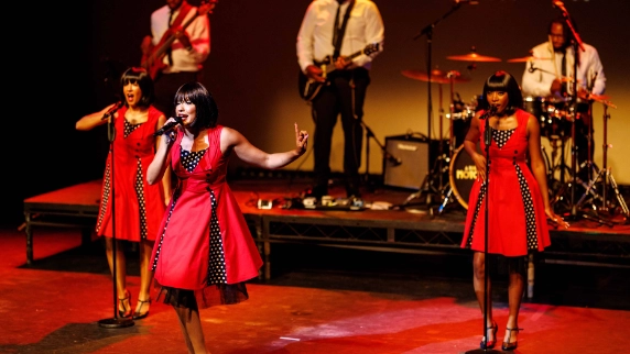 3 female motown performers in red dresses performing alongside a band