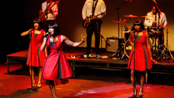 3 female motown performers in red dresses performing alongside a band