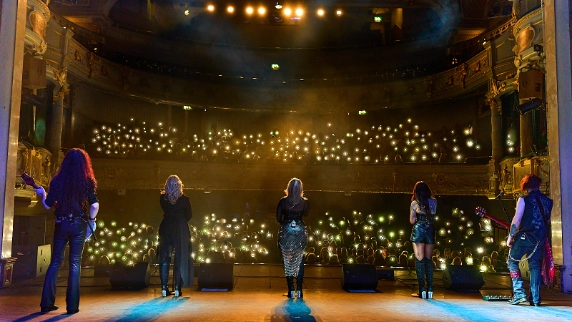 5 ladies performing to an audience using their phones as lights singing onto the stage