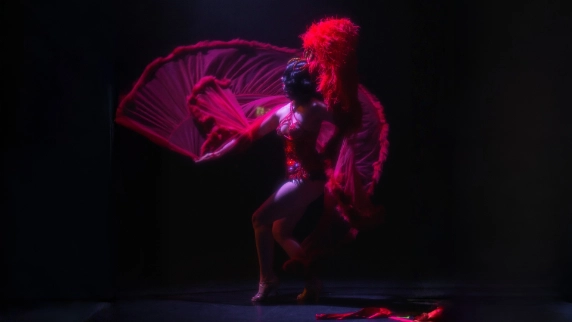 A performer in a dynamic pose is dressed in an elaborate red costume with large feathers, set against a dark background, conveying boldness and drama.