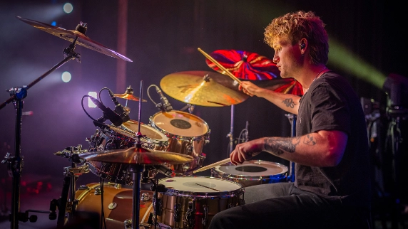 A drummer with curly blonde hair and tattoos performing on stage with dramatic concert lighting in purple, blue, and green tones, creating that energetic live performance atmosphere.