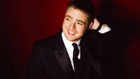Tom Ball wearing a black suit smiling against red background.