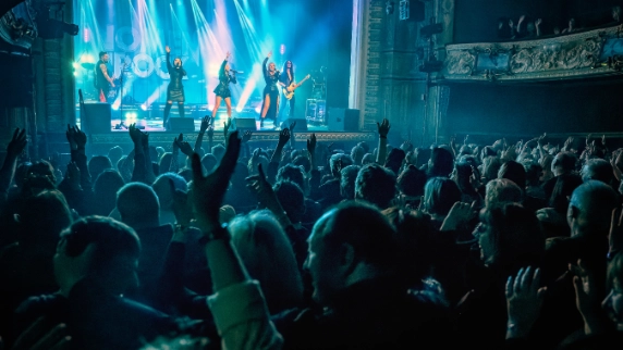 a band consisting of 3 singing ladies, 2 guitarists, a keyboard player and a drummer performing to a crowd of people in a theatre setting