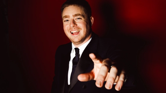 Tom Ball wearing a classic black suit with a white shirt and black tie, photographed against a rich, deep red background.