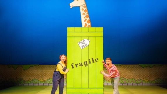 Two playful performers pose with a large green crate labeled “fragile” and “from the zoo,” from which a cartoon giraffe head emerges. The scene is colorful and whimsical.