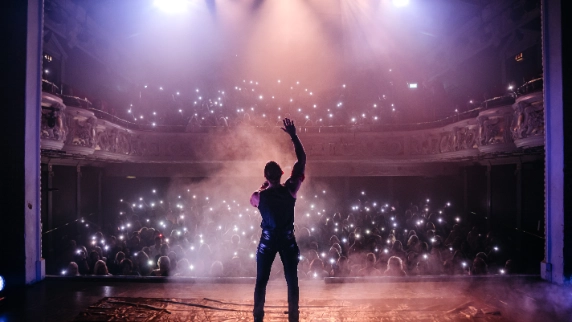 A male performer stands on stage facing a packed theatre audience, raising one hand in the air as he addresses the crowd. Bright stage lights beam down through light haze, while the audience holds up phones with flashlights, creating a sea of glowing lights across the auditorium and balcony.