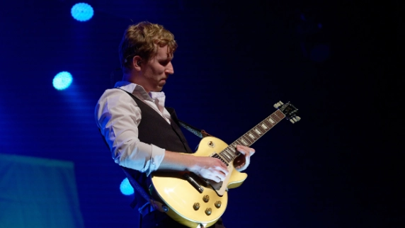 Guitarist in white shirt and vest playing cream-coloured electric guitar on stage with blue lighting