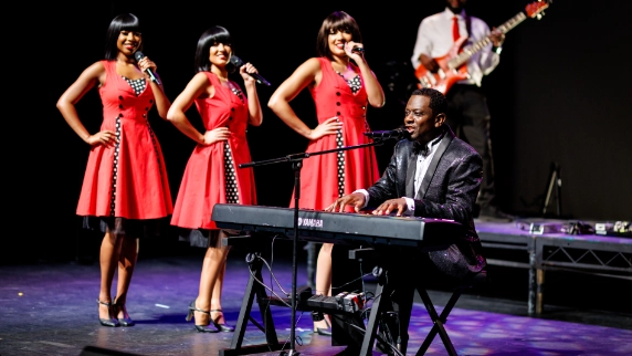 3 female motown performers singing in red dresses alongside a male motown singer sat down playing a keyboard