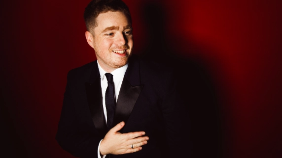 Tom Ball wearing a black suit smiling against red background.