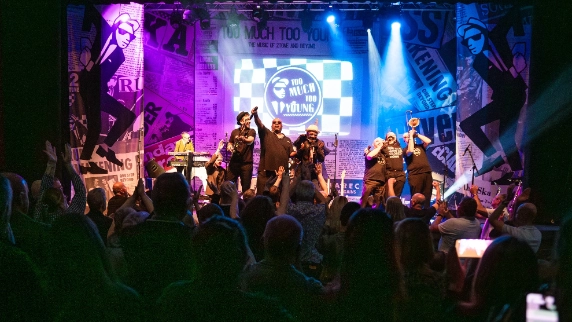 Colour photo of Too Much Too Young performing live on stage in a packed venue. Multiple vocalists stand at the front, raising their arms and engaging the crowd, while brass players and a keyboardist perform behind them.