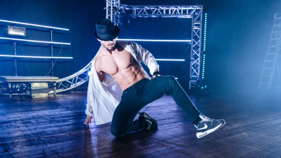 A shirtless male dancer wearing an open white shirt, black pants, sneakers, and a black fedora performs a floor move on a stage.