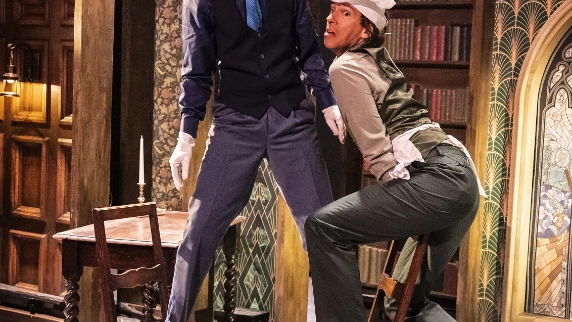 Two actors balancing on chairs set in a library scene on stage 