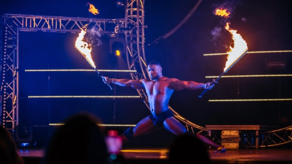 A shirtless performer in black shorts is powerfully balancing with legs bent, wielding flaming batons, under dramatic stage lighting and rigging.